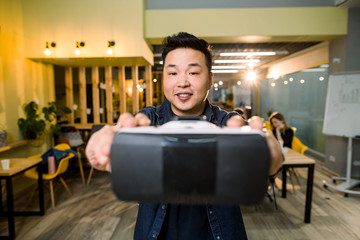 An Asian man holding virtual reality goggle on background of modern coworking studio office, his colleagues sitting at the table on the background. © sofiko14