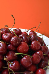 White bowl of fresh cherries on a wooden table on orange background.