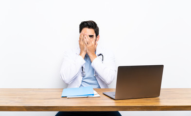 Young doctor man with his laptop over isolated wall covering eyes and looking through fingers