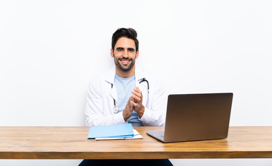 Young doctor man with his laptop over isolated wall applauding