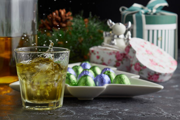 Glass of whiskey with ice and spray on the background of Christmas gifts in holiday boxes, Christmas wreath and candy on a plate in the shape of a Christmas tree.