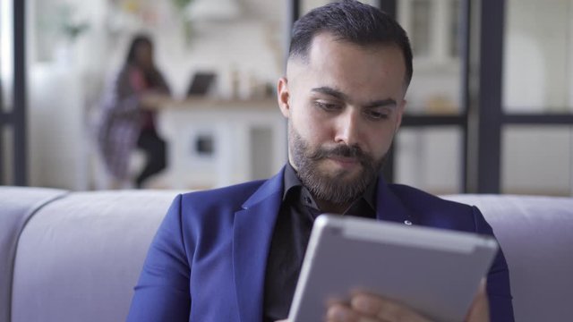 Smiling bearded man in blue suit sitting at home working with his tablet. Leisure indoors. The businessman resting after work. Gadget addiction.