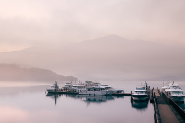 Fototapeta premium Beautiful tranquil landscape at Sun Moon lake in Nantao, Taiwan. Pier with boats and background of foggy mountains. Concept of peaceful, traanquility of nature.
