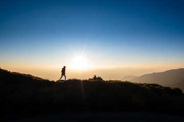 Silhoutte of woman walking on top of the hill with background of sunray during sunset. Concept of hiker, discover, freedom.