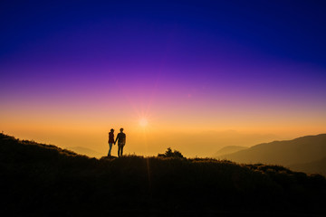 Silhoutte of a couple hold hand of each other looking at sunset on top of hill with background of mountain and colorful color of sunset. Concept of teamwork, successful.