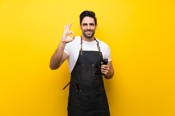 Young hairdresser man over isolated yellow background showing ok sign with fingers