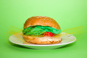 .Fresh tasty burger with plastic waste inside on white plate on green background.  Recycled waste in our food concept. Plastic pollution