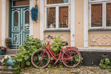 A red retro bike on a cobblestone pavement, leaning aganst a wall with hollyhochs  in Ærøskøbing