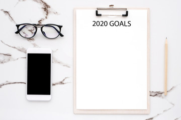 2020 goals on blank note paper with copy space for text and smart phone with blank screen on white marble background, new year aim to success in business background