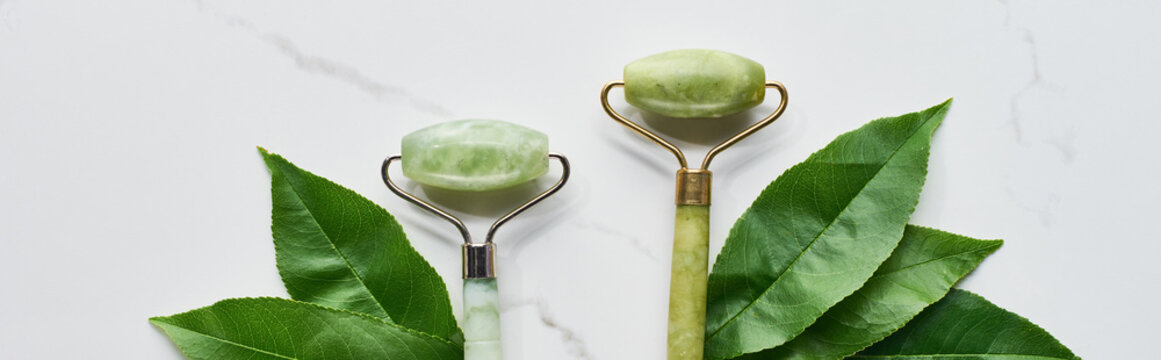 panoramic shot of jadeite face rollers with plant leaves on marble surface
