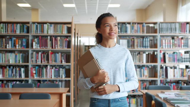 Cheerful teenage girl in casual clothing is walking with books in high school library smiling looking around. Modern education, people and lifestyle concept.