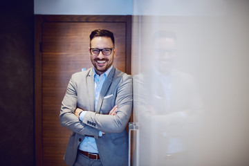 Handsome bearded caucasian businessman with eyeglasses standing in a hall of his apartment with arms crossed