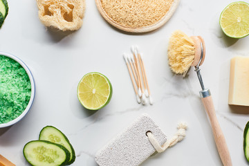 top view of green bath salts, body brush, loofah, bath sponge and pumice stone near fresh lime and cucumber on marble surface