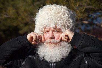 Senior caucasian man in white ethnic sheepskin hat fixing his moustache. Closeup sunny portrait of elderly stylish person in black flight (bomber) jacket at nature against dark forest background.