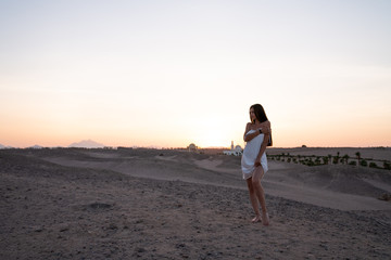 Young beautiful girl in white dress standing among the desert, sunset on the background