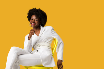 cheerful african american woman sitting on yellow chair and pointing with finger isolated on orange