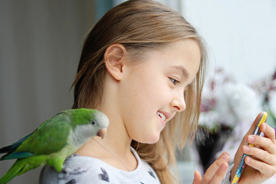 Cute smiling girl with her pet green Monk Parakeet parrot sitting on her shoulder playing game on smartphone. Quaker parrot bird owner. Exotic pet. Pets vs gadgets. Selective focus on girl.