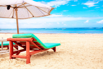A sun lounger under an umbrella on the sandy beach by the ocean and cloudy sky. Vacation background. Idyllic beach landscape.