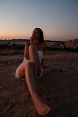 Beautiful dreaming brunette girl in white dress sitting with closed eyes in front of the sunset among the desert