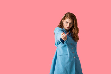 displeased redhead girl looking at camera and showing middle finger isolated on pink