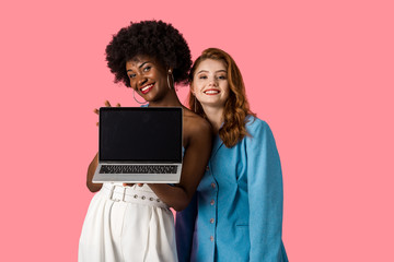 Obraz na płótnie Canvas cheerful multicultural girls smiling near laptop with blank screen isolated on pink