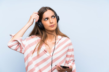 Young woman listening music with a mobile over isolated blue wall having doubts and with confuse face expression