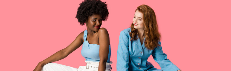 panoramic shot of cheerful multicultural women looking at each other isolated on pink