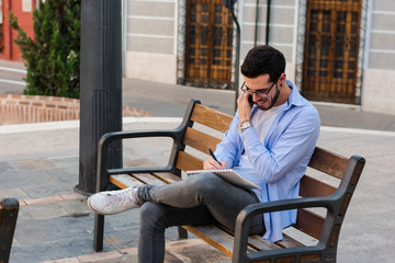 Young businessman is sitting on a bench while he is talking on the mobile and is writing in his notebook