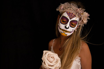 Young blonde and pretty girl, wearing makeup with Mexican Halloween makeup. She is posing in a white dress, a wreath and a flower in her hand