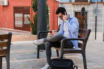 Young businessman is sitting on a bench while he is talking on the mobile and is writing in his notebook