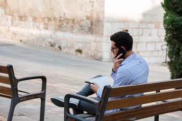 Young businessman is sitting on a bench while he is talking on the mobile and is writing in his notebook