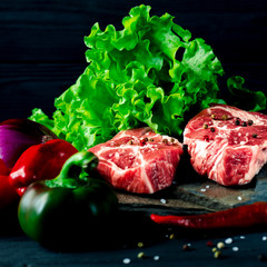 Two juicy steaks lie on a wild stone next to red and green chili peppers and a fresh green salad on a black wooden table