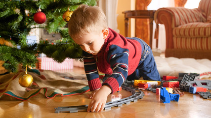 Portrait of little toddler boy building railway and playing with toy train under Christmas tree