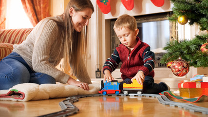 Portrait of boy with his mother playing on floor with toy train and railways. Child receiving presents and toys on New Year or Xmas