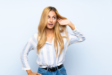 Young blonde woman over isolated blue background listening to something by putting hand on the ear