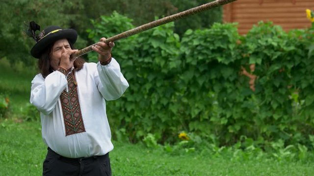 Senior Carpathian man plays trembita instrument. Mature hutsul in traditinal Ukrainian clothing playing trembita. The longest musical instrument.