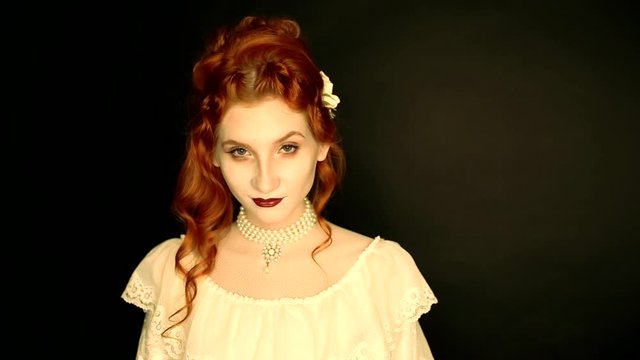 Young vampire woman with long curly hair, pale skin in a white dress on a black background. A beautiful redhead model with red lips. Outfit for halloween. Vampire with open mouth and fangs