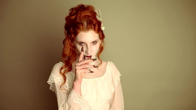 Young vampire woman with long curly hair, pale skin in a white dress on a black background. A beautiful redhead model with red lips. Outfit for halloween. Vampire with open mouth and fangs