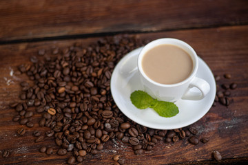 Cup of coffee on a dark background. White milkman. Milk is pouring from it into a cup. On a saucer there are mint leaves, brown grains are scattered nearby, cupcakes cooked at home lie. 
