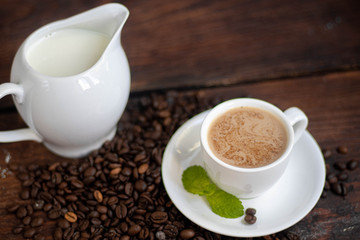 Cup of coffee on a dark background. White milkman. Milk is pouring from it into a cup. On a saucer there are mint leaves, brown grains are scattered nearby, cupcakes cooked at home lie. 