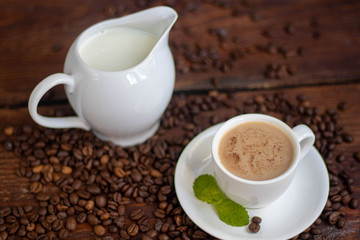 Cup of coffee on a dark background. White milkman. Milk is pouring from it into a cup. On a saucer there are mint leaves, brown grains are scattered nearby, cupcakes cooked at home lie. 