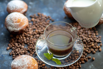 Cup of coffee on a dark background. White milkman. Milk is pouring from it into a cup. On a saucer there are mint leaves, brown grains are scattered nearby, cupcakes cooked at home lie. 