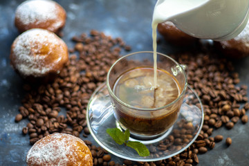 Cup of coffee on a dark background. White milkman. Milk is pouring from it into a cup. On a saucer there are mint leaves, brown grains are scattered nearby, cupcakes cooked at home lie. 