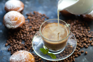 Cup of coffee on a dark background. White milkman. Milk is pouring from it into a cup. On a saucer there are mint leaves, brown grains are scattered nearby, cupcakes cooked at home lie. 