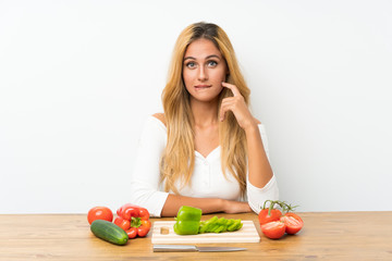 Young blonde woman with vegetables in a table having doubts and with confuse face expression