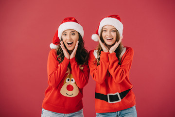 attractive and smiling women in sweaters and santa hats looking at camera isolated on red