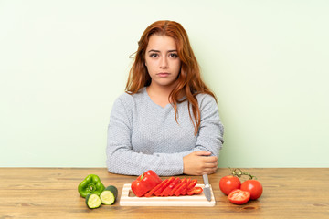 Teenager redhead girl with vegetables in a table keeping arms crossed