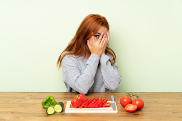 Teenager redhead girl with vegetables in a table covering eyes and looking through fingers