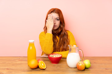 Teenager redhead girl having breakfast in a table covering a eye by hand