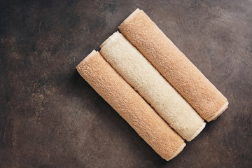 Three terry beige towels rolled up on a dark brown rustic background. Top view, flat lay, copy space.
